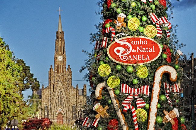 Pinheiro de Natal com uma placa pendurada escrita "Sonho de Natal de Canela" e ao fundo a Catedral de Pedra